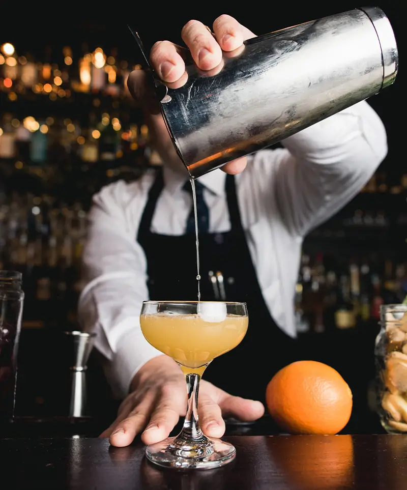 Bartender pouring a handcrafted cocktail into a coupe glass with citrus garnish and bar tools visible in the scene